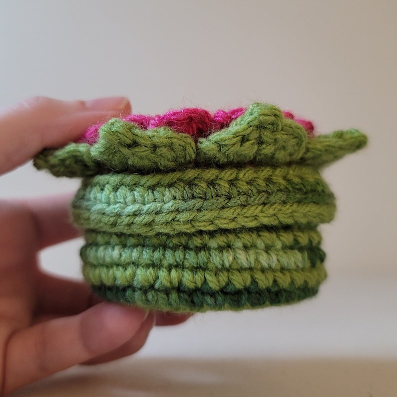 May include: A crocheted flower pot with a pink flower on top. The pot is made of green yarn and has a layered design.