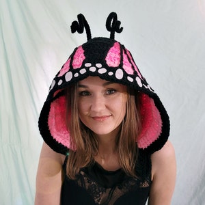 May include: A black and pink crochet butterfly hood with white accents. The hood features large, wing-shaped panels in black with pink and white detailing. Two black antennae are attached to the top of the hood.