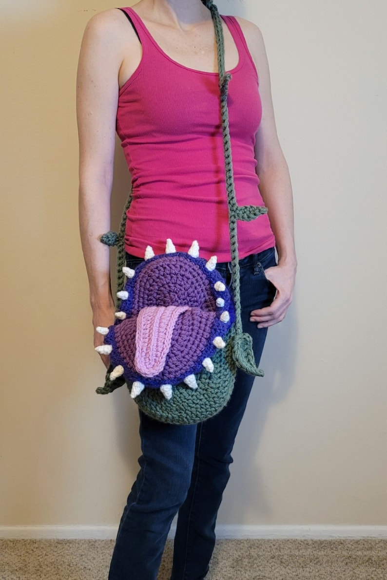 May include: A green, purple, and pink crocheted bag shaped like a Venus flytrap with a white crocheted tongue. The bag is worn as a crossbody bag.