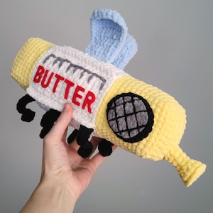 Op de afbeelding: Een handgemaakt, pluche speelgoed in de vorm van een pakje boter, met een gele body en witte verpakking. Het woord "BUTTER" staat in rode letters op de verpakking. Het heeft zwarte poten, een grijs en zwart oog en blauwe vleugels.