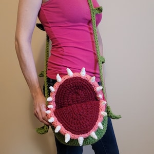 May include: A green crocheted bag shaped like a plant with a red and pink mouth. The bag is worn as a crossbody bag.