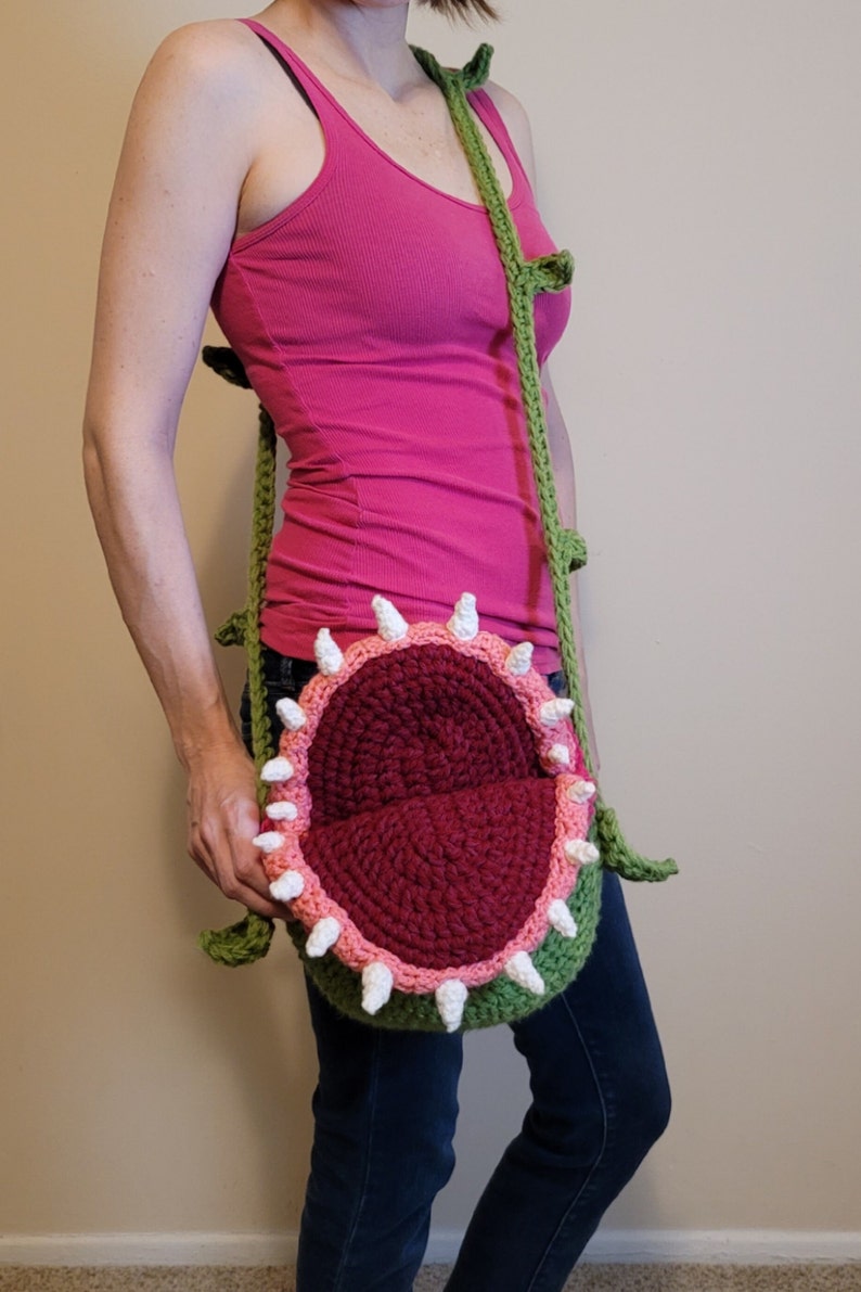 May include: A green crocheted bag shaped like a plant with a red and pink mouth. The bag is worn as a crossbody bag.