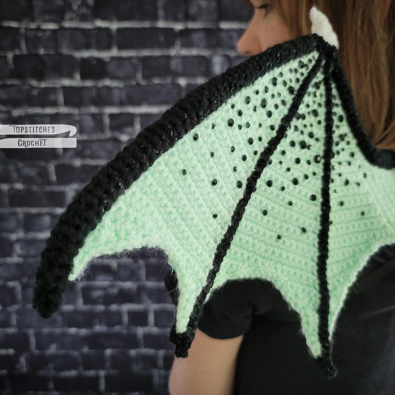 K&ouml;nnte beinhalten: Geh&auml;kelter Drachenfl&uuml;gel in Mintgr&uuml;n mit schwarzem Rand und schwarzen Perlenakzenten. Der Fl&uuml;gel wird von einer Person gehalten. Der Text "TOPSTITCHES CROCHET" ist auf der linken Seite des Bildes zu sehen.