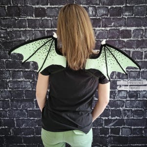 K&ouml;nnte beinhalten: Geh&auml;kelte Drachenfl&uuml;gel in Hellgr&uuml;n und Schwarz, &uuml;ber den Schultern getragen. Die Fl&uuml;gel haben ein detailliertes Muster und sind an schwarzen Tr&auml;gern befestigt. Die Person tr&auml;gt ein schwarzes T-Shirt und eine gr&uuml;ne Hose. Der Hintergrund ist eine dunkle Backsteinmauer.