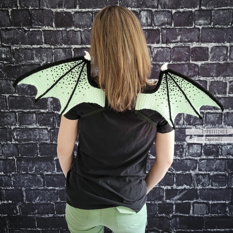 K&ouml;nnte beinhalten: Geh&auml;kelte Drachenfl&uuml;gel in Hellgr&uuml;n und Schwarz, &uuml;ber den Schultern getragen. Die Fl&uuml;gel haben ein detailliertes Muster und sind an schwarzen Tr&auml;gern befestigt. Die Person tr&auml;gt ein schwarzes T-Shirt und eine gr&uuml;ne Hose. Der Hintergrund ist eine dunkle Backsteinmauer.