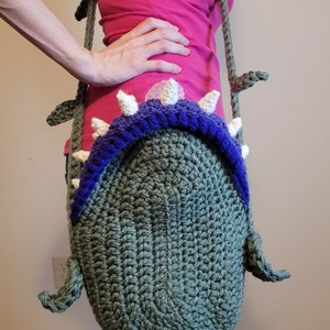 May include: A green crocheted bag shaped like a monster with white teeth and a purple rim. The bag is being worn as a shoulder bag.