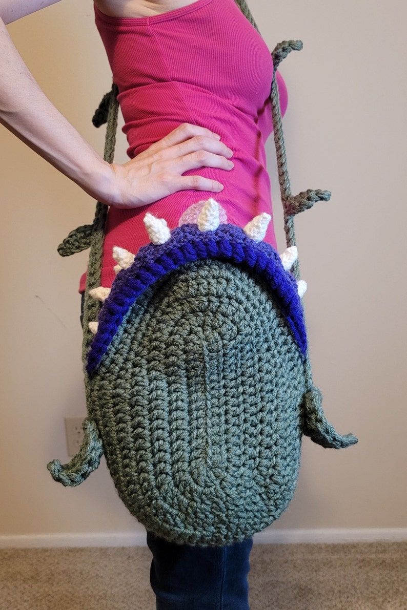 May include: A green crocheted bag shaped like a monster with white teeth and a purple rim. The bag is being worn as a shoulder bag.