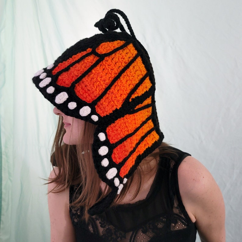 May include: A crocheted butterfly wing hat in black, orange, and white. The hat features a monarch butterfly wing design with black outlines, orange filling, and white spots. The hat is worn by a person.