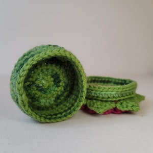 May include: Two crocheted green and pink flower-shaped baskets. The baskets are made of yarn and have a textured surface.