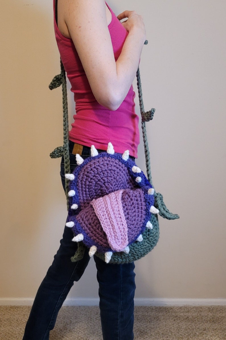 May include: A crocheted shoulder bag shaped like a Venus flytrap with a purple and pink mouth and green vines.