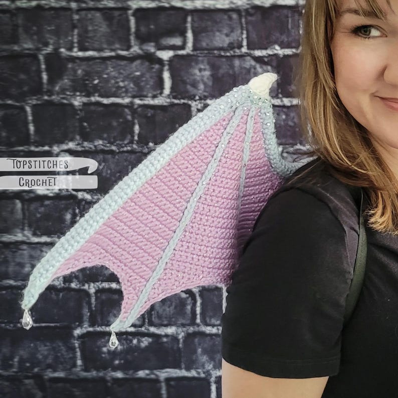 K&ouml;nnte beinhalten: Geh&auml;kelte Drachenfl&uuml;gel in Pastellrosa und Hellblau, mit silbernen Akzenten und transparenten, tropfenf&ouml;rmigen Verzierungen. Die Fl&uuml;gel werden auf dem R&uuml;cken getragen, mit dem Text "Topstitches Crochet" sichtbar.