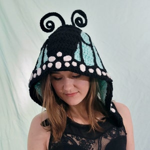 May include: A black, turquoise, and white crochet butterfly hood. The hood features a black top with two curled antennae. The turquoise wings are outlined in black and have white polka dots. The hood is worn by a person.