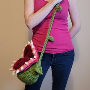 May include: A crocheted green, pink, and white plant-shaped bag with a long green strap. The bag is shaped like a Venus flytrap with a large open mouth.
