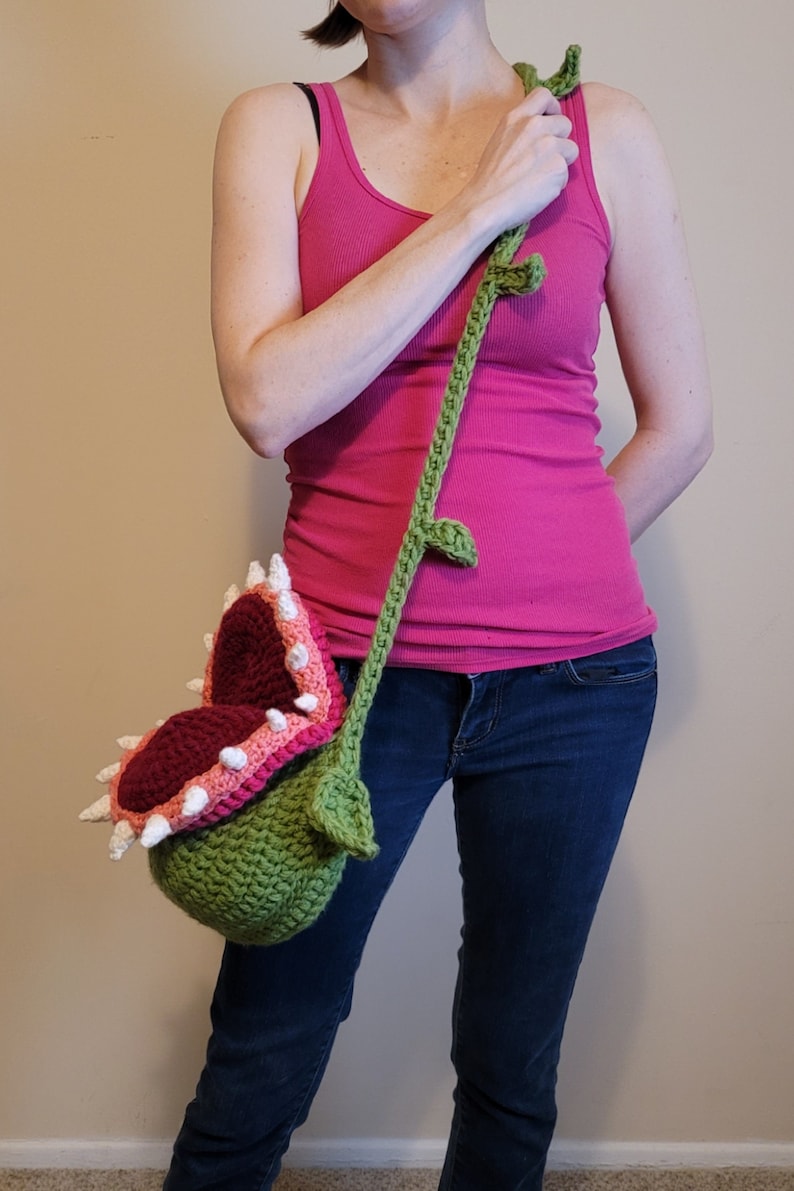 May include: A crocheted green, pink, and white plant-shaped bag with a long green strap. The bag is shaped like a Venus flytrap with a large open mouth.
