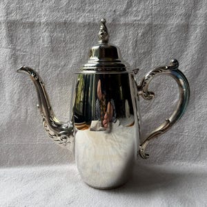 May include: A polished silver-toned coffee pot featuring a tall, cylindrical body, a curved spout with ornate embossed detailing, a matching scrolled handle, and a removable lid topped with a decorative finial. The surface is highly reflective.