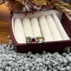 Könnte beinhalten: Ein goldener Ring mit einem roten Edelstein und zwei kleineren weißen Edelsteinen. Der Ring befindet sich in einer roten Samt-Ringbox.