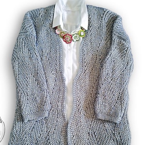 May include: A gray knitted cardigan with a cable knit pattern. The cardigan is open in the front and has long sleeves. A white shirt is worn underneath the cardigan, and a floral necklace is worn around the neck.
