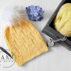 Pode incluir: Um gorro de malha amarelo com um pompom branco. O gorro tem um padrão de malha trançada e está sobre uma superfície de madeira branca. Há um novelo de fio amarelo e um novelo de fio azul numa caixa de madeira no fundo.