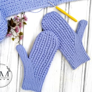 May include: A pair of lavender crocheted mittens with a textured stitch pattern. The mittens are laid out on a white wooden surface with a ball of yarn and a crochet hook.