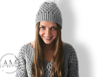 May include: A grey knitted beanie with a white pom pom. The beanie has a lacy pattern and is being worn by a person with long brown hair.