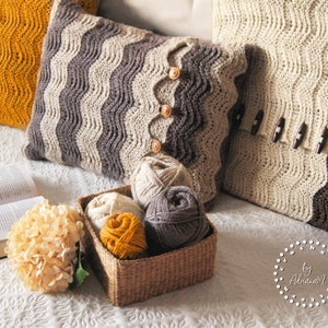 May include: Three crocheted pillows with a wavy pattern in beige, brown, and mustard yellow. The pillows are decorated with buttons and are arranged on a white bedspread with a book and a bouquet of white flowers.