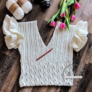 May include: Cream-colored, handmade crochet top with a V-neck and ruffled sleeves. The top features a textured stitch pattern on the lower portion and a red crochet hook. Two balls of white yarn and pink tulips are also visible.
