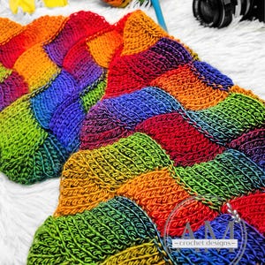 Könnte beinhalten: Eine handgefertigte, gehäkelte Decke in Regenbogenfarben. Die Decke hat ein strukturiertes Design mit Quadraten in Rot, Orange, Gelb, Grün, Blau und Lila. Ein Knäuel Regenbogenwolle, eine Häkelnadel und eine Kamera sind ebenfalls zu sehen.