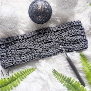 May include: A gray crochet headband with a cable knit pattern. The headband is lying on a white fluffy surface with a crochet hook next to it.