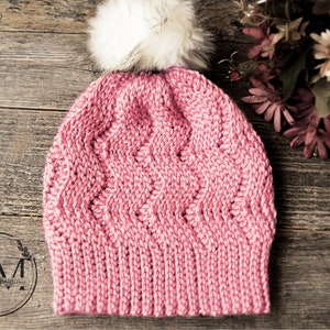 Puede incluir: Un gorro de punto rosa con un pompón de piel sintética blanca. El gorro tiene un patrón de punto texturizado y está fotografiado sobre una superficie de madera.