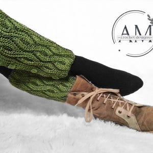 May include: Green knitted leg warmers with buttons, worn over black leggings and brown boots. The leg warmers have a cable knit pattern. The image includes the text "AM -crochet designs-"