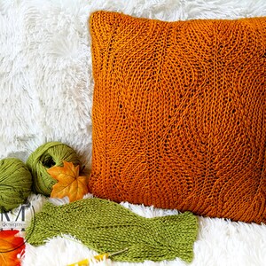 May include: A close-up of a knitted orange pillow with a leaf pattern. The pillow is on a white fluffy surface with two balls of green yarn and a fall leaf.