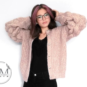 Puede incluir: Un cárdigan de punto rosa con cierre de botones y mangas grandes y abullonadas. El cárdigan está hecho con un hilo suave y texturizado.