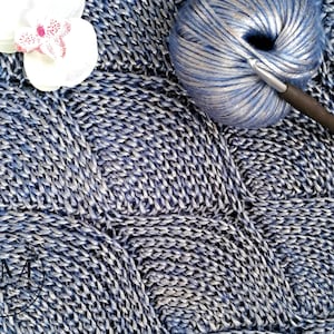 Puede incluir: Primer plano de un hilo azul y plateado con un ganchillo. El hilo está en una bola y el ganchillo está encima. El hilo está sobre una tela de punto azul y plateado.