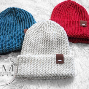 May include: Three crocheted beanies in different colors: teal, white with flecks of gold, and red. Each beanie has a brown leather patch with a white stitch detail.