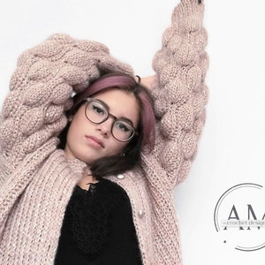 Puede incluir: Un cárdigan de punto rosa con mangas grandes y abullonadas. El cárdigan tiene cierre de botones y un brillo sutil. La modelo lleva el cárdigan con un top negro debajo.