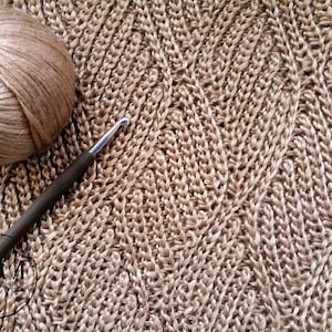 Może przedstawiać: Zbliżenie na tkaninę robioną na szydełku z kłębkiem brązowej włóczki i szydełkiem. Tkanina ma fakturowany, falisty wzór.