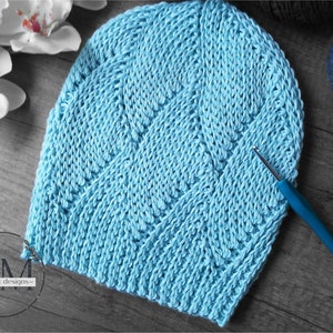 May include: A light blue, handmade crochet beanie with a textured chevron pattern. A crochet hook with a blue handle rests on the hat. The hat is displayed on a wooden surface, with white flowers and a ball of yarn in the background.
