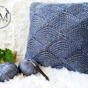 May include: A blue and silver yarn pillow with a wave pattern. The pillow is on a white fluffy surface with two balls of yarn and a crochet hook.