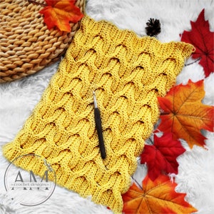 May include: A yellow crocheted blanket with a wavy pattern. The blanket is on a white fuzzy surface with fall leaves.