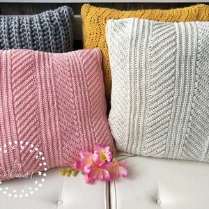 May include: Four crocheted throw pillows in different colors and patterns. The pillows are arranged on a white couch. The pillows are gray, pink, yellow, and white.