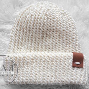 May include: A white crocheted beanie with a brown leather patch that says "AM - crochet designs -" on the side.