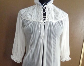 May include: A white sheer pleated robe featuring a high ruffled collar with a tie closure. The garment has long sleeves with ruffled cuffs and a delicate lace yoke panel across the upper chest and shoulders, creating a layered texture.