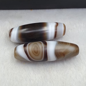 May include: Two cylindrical stone beads with smooth, polished surfaces. The top bead features dark brown and white horizontal bands. The bottom bead displays a prominent brown circular eye pattern surrounded by white and light brown concentric rings.