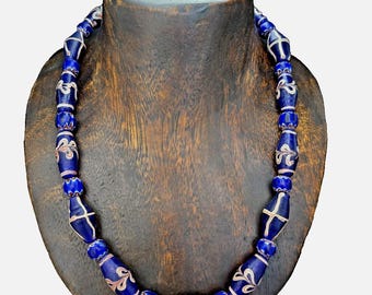 May include: A necklace with dark blue glass beads of varying shapes and sizes. The beads have white and red floral designs and are strung together. The necklace is displayed on a wooden bust.