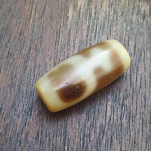 May include: A cylindrical, cream-colored bead with brown, abstract markings. The bead has rounded ends and appears to be made of stone or a similar material. The bead is lying on a wooden surface.