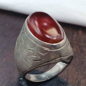 May include: A silver-toned metal ring featuring a large, smooth, oval-shaped deep red stone set in the center. The band of the ring displays intricate, etched floral patterns that wrap around the sides, providing a textured contrast to the polished stone.