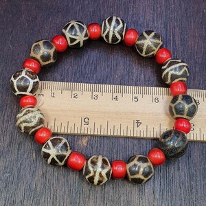 Può includere: Un braccialetto di perline con perline scure decorate e piccole perline rosse rotonde. Le perline decorate hanno uno sfondo chiaro con disegni geometrici marroni scuri. Il braccialetto è lungo circa 15 cm.