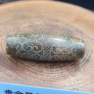 Puede incluir: Una cuenta de jade verde con intrincados patrones tallados de líneas en espiral. La cuenta tiene forma ovalada y una superficie lisa y pulida.