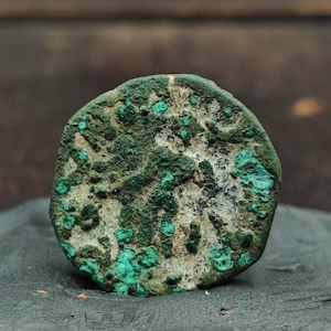 May include: A close-up of an old, green and white coin with a design on the front. The coin is worn and has a patina.