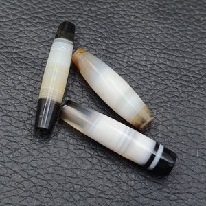 Könnte beinhalten: Drei weiße und schwarze gebänderte Achatperlen, auch bekannt als Dzi-Perlen, mit braunen und weißen Streifen. Die Perlen sind oval geformt und haben eine glatte, polierte Oberfläche.